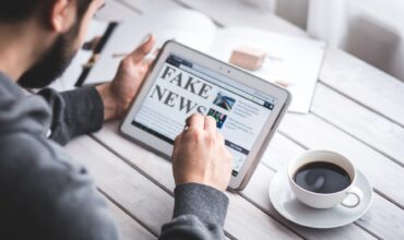Un homme sur une tablette lit de l'information. Le contenu de l'écran indique fake news. Une tasse de café est posée à côté.