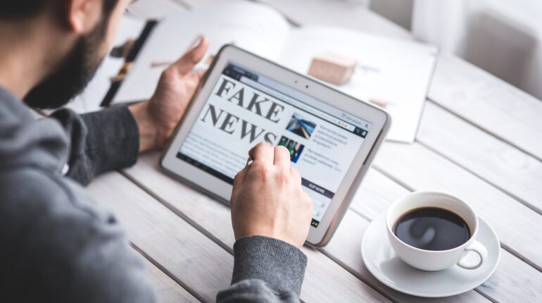 Un homme sur une tablette lit de l'information. Le contenu de l'écran indique fake news. Une tasse de café est posée à côté.